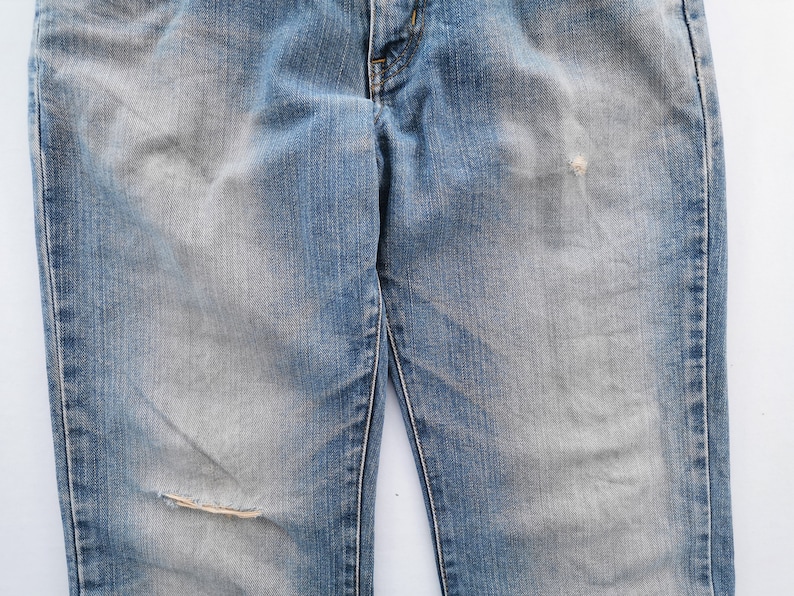 May include: Faded blue denim jeans with a distressed look. The jeans have a worn appearance with a ripped knee and faded patches. The classic five-pocket design is visible.