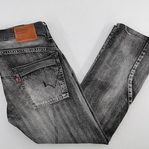 May include: A pair of faded black denim jeans. The jeans feature a classic five-pocket design, a leather patch, and a red tag. The jeans are folded, showcasing the details of the wash and stitching.