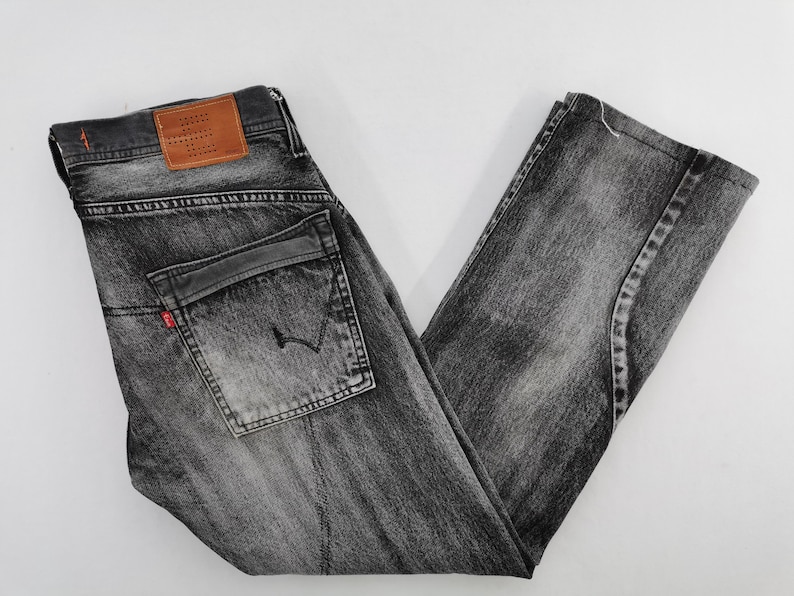 May include: A pair of faded black denim jeans. The jeans feature a classic five-pocket design, a leather patch, and a red tag. The jeans are folded, showcasing the details of the wash and stitching.