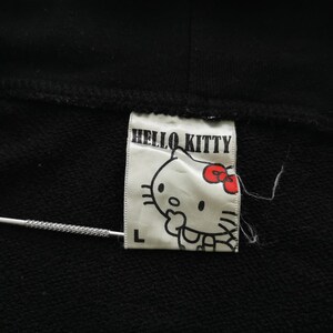 May include: A black garment with a white label displaying "HELLO KITTY" and a cartoon image of Hello Kitty with a red bow. The label also has the letter "L". A silver needle is also in the image.
