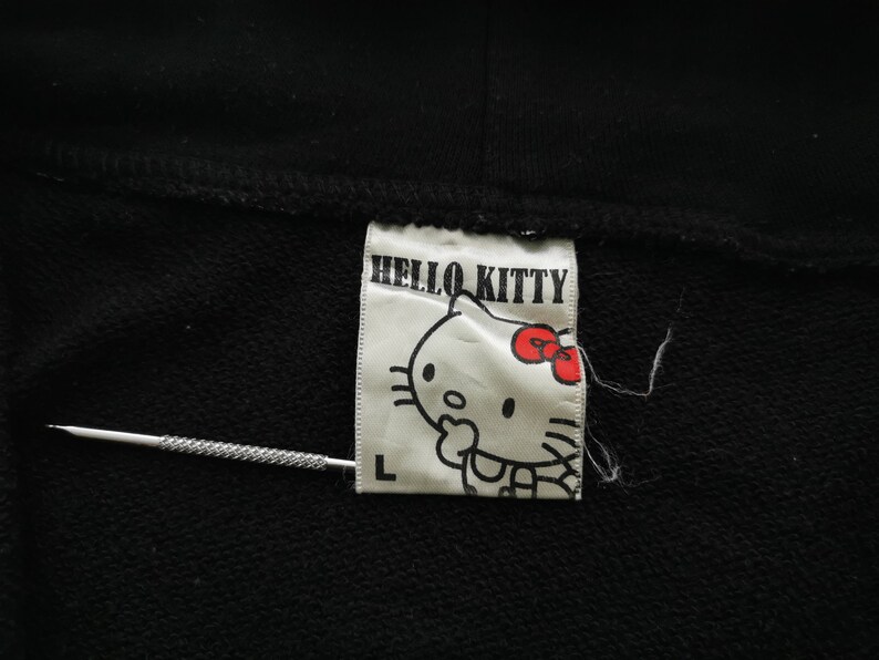 May include: A black garment with a white label displaying "HELLO KITTY" and a cartoon image of Hello Kitty with a red bow. The label also has the letter "L". A silver needle is also in the image.
