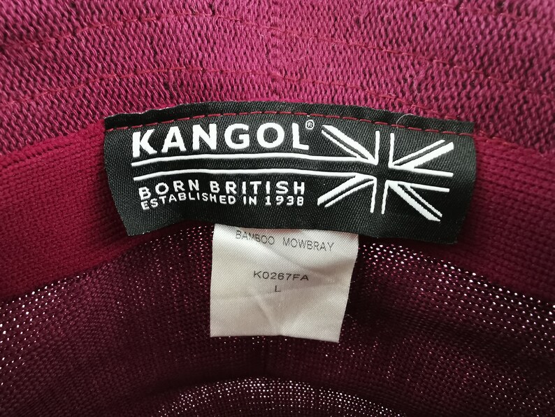 K&ouml;nnte beinhalten: Nahaufnahme des Etiketts eines bordeauxfarbenen Kangol-Hutes. Das Etikett lautet "KANGOL" &uuml;ber "BORN BRITISH ESTABLISHED IN 1938" mit einer Union-Jack-Grafik. Darunter steht "BAMBOO MOWBRAY" und "K0267FA L".