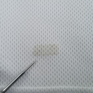 May include: Close-up of a white, textured fabric with a small, dark stain. The fabric has a perforated pattern, and a silver tool is in the foreground, pointing towards the stain. The fabric appears to be a sports jersey material.