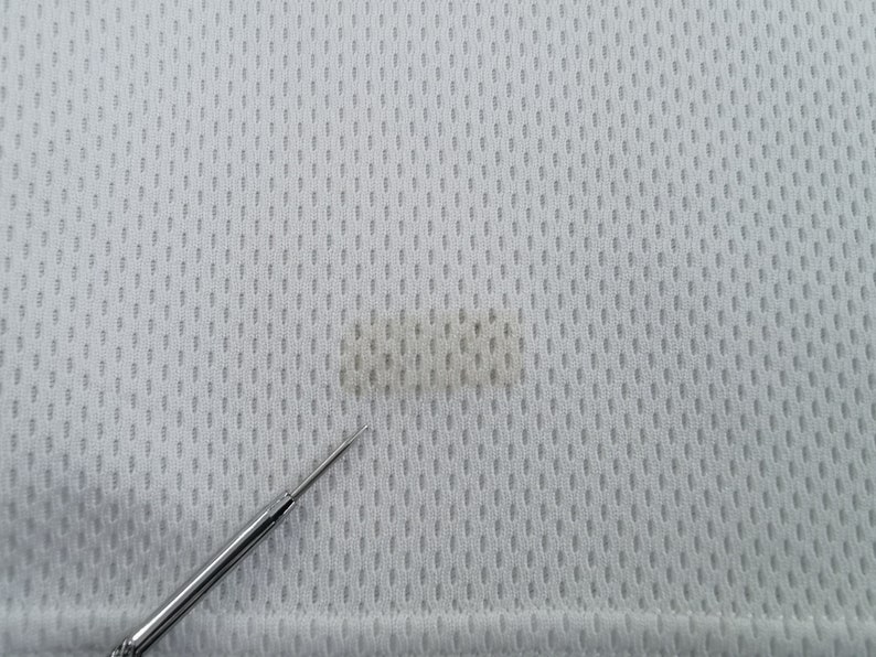 May include: Close-up of a white, textured fabric with a small, dark stain. The fabric has a perforated pattern, and a silver tool is in the foreground, pointing towards the stain. The fabric appears to be a sports jersey material.