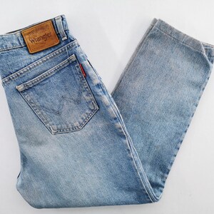 Op de afbeelding: Lichtblauwe Wrangler-jeans met een klassiek ontwerp met vijf zakken. De jeans hebben een bruine leren patch met het Wrangler-logo op de achterkant en een rood label op de achterzak. De denim is vervaagd, waardoor het een vintage uitstraling heeft.