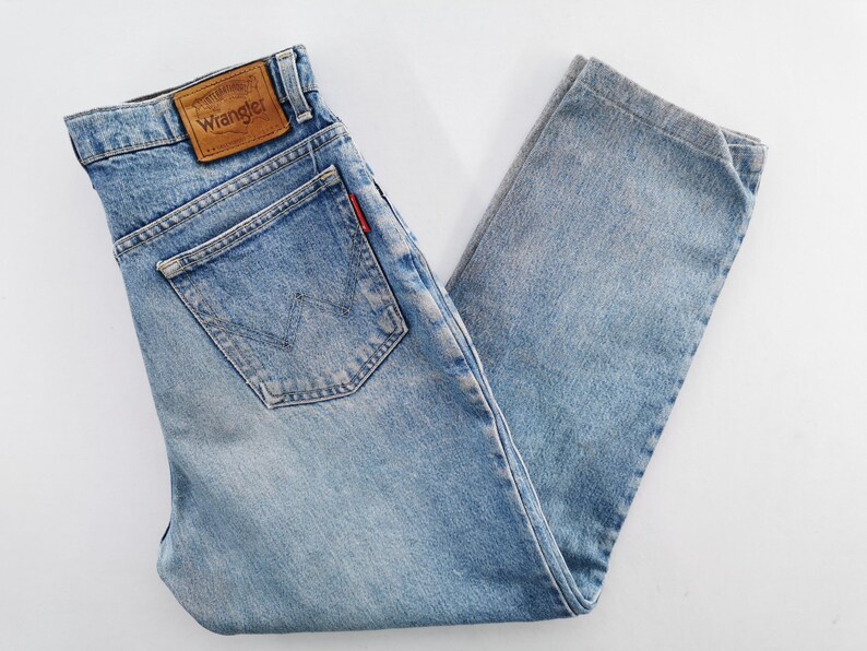 Op de afbeelding: Lichtblauwe Wrangler-jeans met een klassiek ontwerp met vijf zakken. De jeans hebben een bruine leren patch met het Wrangler-logo op de achterkant en een rood label op de achterzak. De denim is vervaagd, waardoor het een vintage uitstraling heeft.