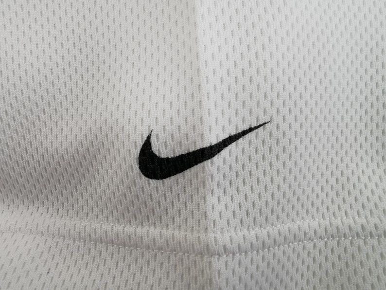 May include: Close-up of white athletic mesh fabric featuring a black Nike swoosh logo. The fabric has a textured, breathable design, and the logo is positioned in the upper right corner.