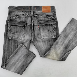 May include: Faded black denim jeans with a classic five-pocket design. Features include two back pockets, a small red tag, and a brown leather patch. The jeans are laid flat on a white surface.
