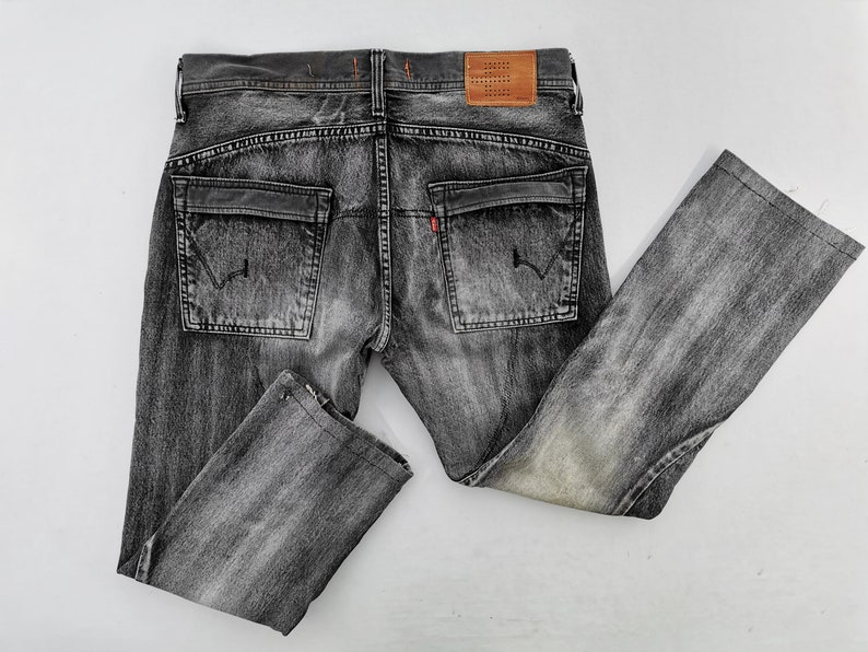May include: Faded black denim jeans with a classic five-pocket design. Features include two back pockets, a small red tag, and a brown leather patch. The jeans are laid flat on a white surface.