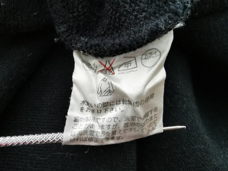 以下が含まれることがあります： 黒い衣服のクローズアップ。白いケアラベルが付いています。ラベルには、アイロン禁止マークやタンブル乾燥禁止マークなど、いくつかのケアシンボルが表示されています。日本語のテキストも表示されています。