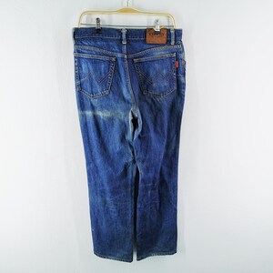 May include: Blue denim jeans with a classic five-pocket design, hanging on a wooden hanger. The jeans have a faded wash and a leather patch with the brand name. A red tag is visible on the back pocket.