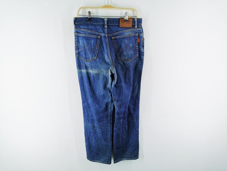 May include: Blue denim jeans with a classic five-pocket design, hanging on a wooden hanger. The jeans have a faded wash and a leather patch with the brand name. A red tag is visible on the back pocket.