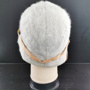 May include: A light gray, fuzzy hat with a rounded crown and a tan-colored band. The hat is displayed on a mannequin head against a dark background. The hat's texture appears soft and plush, suggesting a warm accessory.