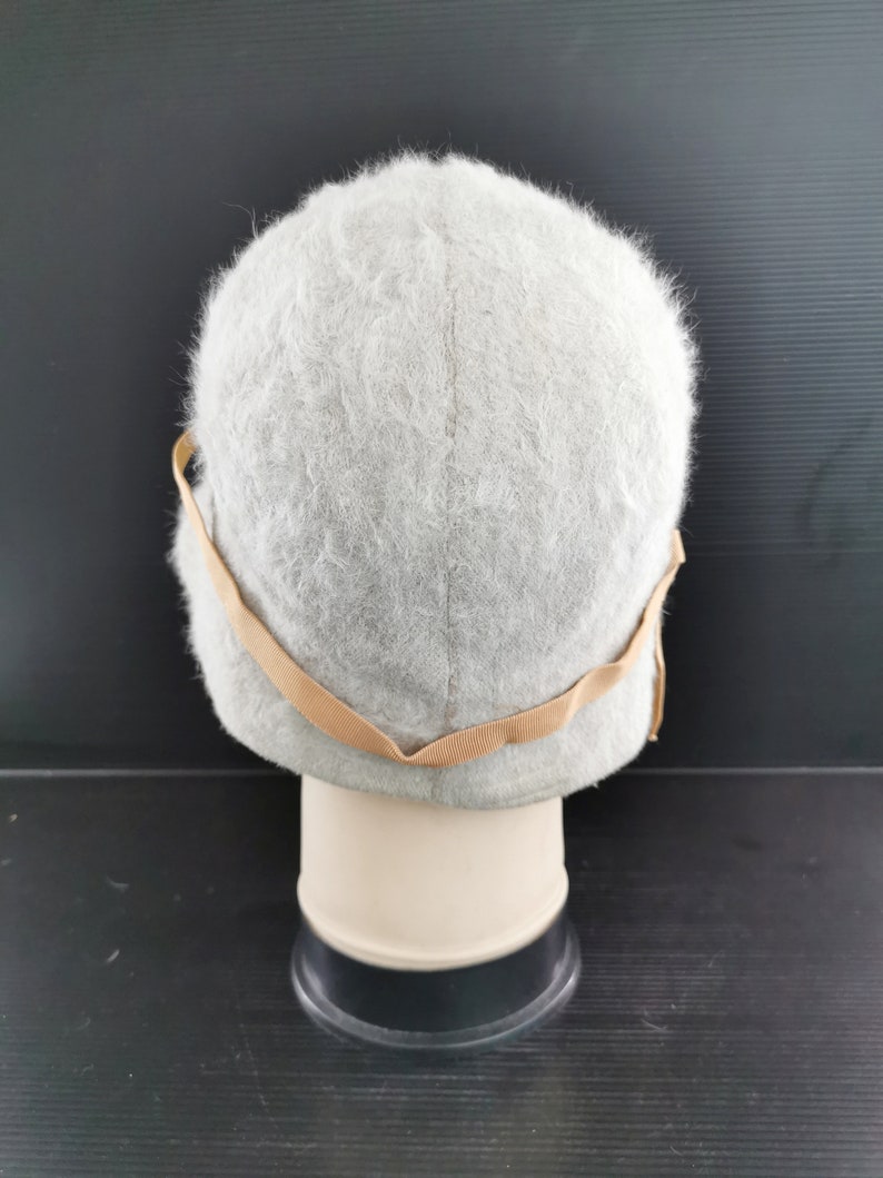 May include: A light gray, fuzzy hat with a rounded crown and a tan-colored band. The hat is displayed on a mannequin head against a dark background. The hat's texture appears soft and plush, suggesting a warm accessory.