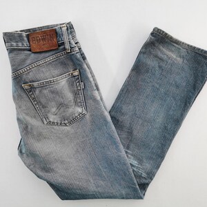 May include: Faded blue denim jeans with a classic five-pocket design. The jeans feature a leather patch with the brand name "EDWIN" on the back waistband. The jeans are folded and laid flat on a white surface.