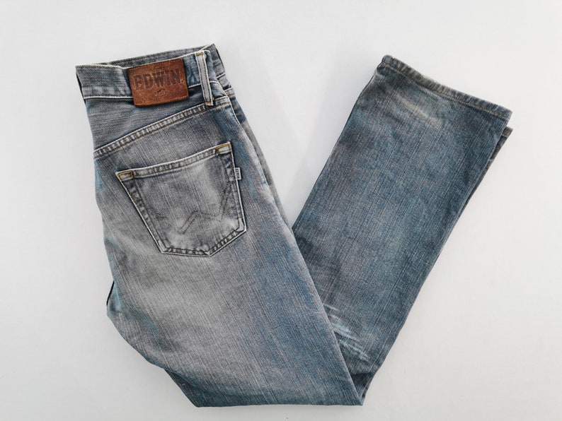 May include: Faded blue denim jeans with a classic five-pocket design. The jeans feature a leather patch with the brand name "EDWIN" on the back waistband. The jeans are folded and laid flat on a white surface.