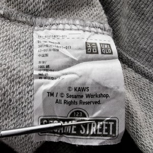 K&ouml;nnte beinhalten: Nahaufnahme eines grauen Stoffetiketts mit Text der Marke Uniqlo. Das Etikett enth&auml;lt das Sesame Street-Logo und das KAWS-Copyright. Das Etikett ist an einem grauen Sweatshirt befestigt.