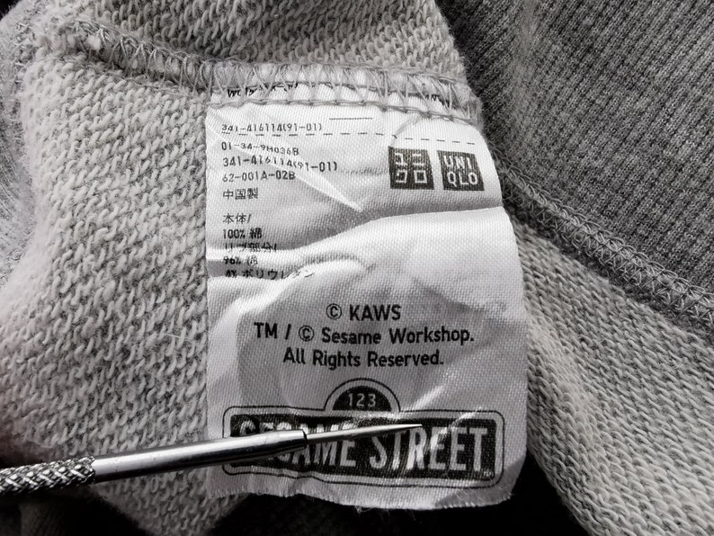 K&ouml;nnte beinhalten: Nahaufnahme eines grauen Stoffetiketts mit Text der Marke Uniqlo. Das Etikett enth&auml;lt das Sesame Street-Logo und das KAWS-Copyright. Das Etikett ist an einem grauen Sweatshirt befestigt.