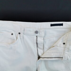 May include: Close-up of off-white denim jeans with a partially unzipped fly. The jeans have a button closure, front pockets, and dark metal rivets. The fabric appears to be a light-weight denim.