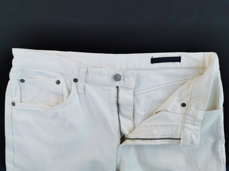 May include: Close-up of off-white denim jeans with a partially unzipped fly. The jeans have a button closure, front pockets, and dark metal rivets. The fabric appears to be a light-weight denim.