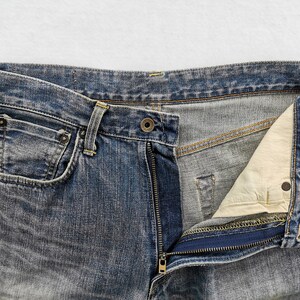 May include: Close-up of blue denim jeans with an open zipper. The jeans have a classic five-pocket design, with visible stitching and a button closure. The denim has a faded, worn appearance.