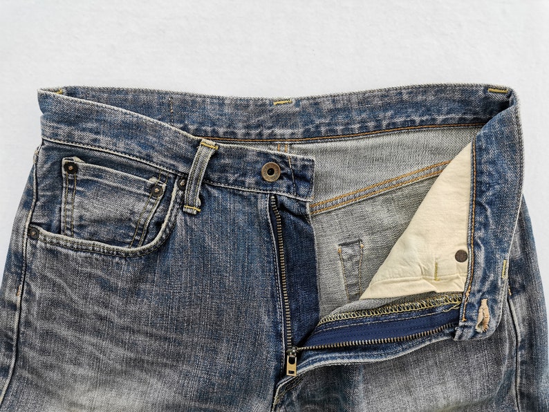 May include: Close-up of blue denim jeans with an open zipper. The jeans have a classic five-pocket design, with visible stitching and a button closure. The denim has a faded, worn appearance.