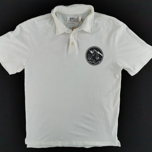 Puede incluir: Polo blanco con mangas cortas y cuello. La camisa presenta un gr&aacute;fico circular negro en el pecho con un rayo y un dise&ntilde;o de olas. La marca "AVIREX LTD" es visible en la etiqueta.
