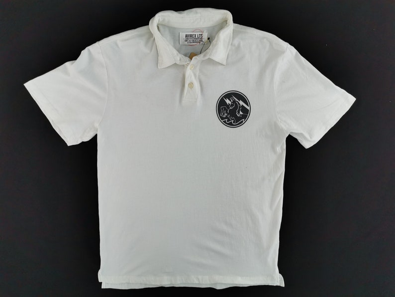Puede incluir: Polo blanco con mangas cortas y cuello. La camisa presenta un gr&aacute;fico circular negro en el pecho con un rayo y un dise&ntilde;o de olas. La marca "AVIREX LTD" es visible en la etiqueta.