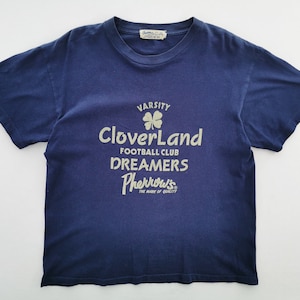 May include: A navy blue t-shirt with a crew neck and short sleeves. The shirt displays the text "Varsity CloverLand Football Club Dreamers Pherrow's The Mark of Quality" and a four-leaf clover graphic.