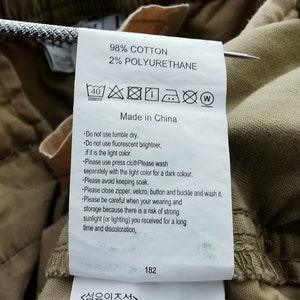 May include: A close-up of a clothing tag with the text "98% COTTON 2% POLYURETHANE Made in China". The tag includes care instructions with symbols and text. The tag is attached to tan fabric.
