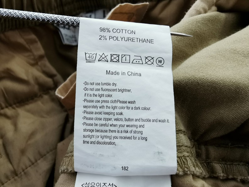 May include: A close-up of a clothing tag with the text "98% COTTON 2% POLYURETHANE Made in China". The tag includes care instructions with symbols and text. The tag is attached to tan fabric.