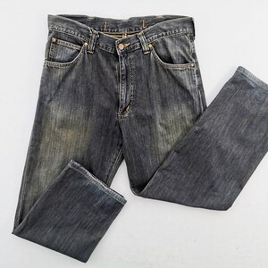 Peut inclure: Un jean en denim noir d&eacute;lav&eacute; avec une conception classique &agrave; cinq poches. Le jean a une fermeture &agrave; bouton et &agrave; glissi&egrave;re, des passants de ceinture et un aspect l&eacute;g&egrave;rement us&eacute;. Le jean est pos&eacute; &agrave; plat sur une surface blanche.