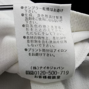 May include: A close-up of a white fabric label with Japanese text detailing care instructions. The label advises against tumble drying, the use of fluorescent detergents, and ironing. It also includes a phone number and company details.