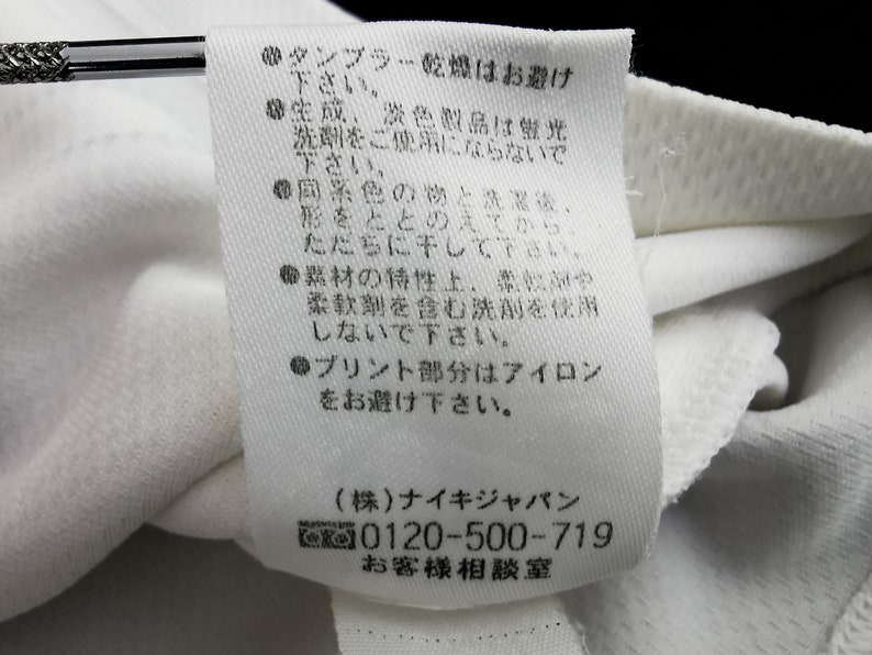 May include: A close-up of a white fabric label with Japanese text detailing care instructions. The label advises against tumble drying, the use of fluorescent detergents, and ironing. It also includes a phone number and company details.
