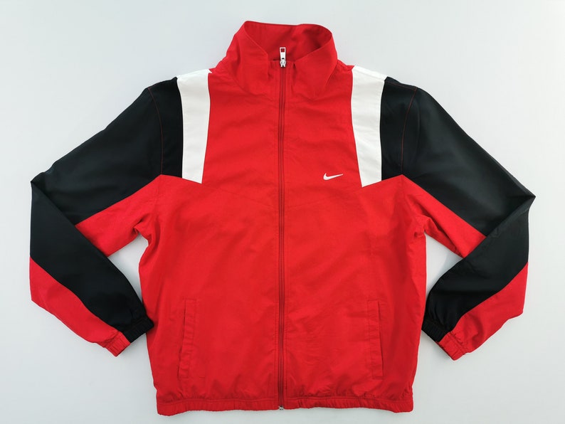 May include: Red, black, and white zip-up track jacket. The jacket features a red body with black sleeves and white accents on the shoulders. A small white Nike logo is on the chest.