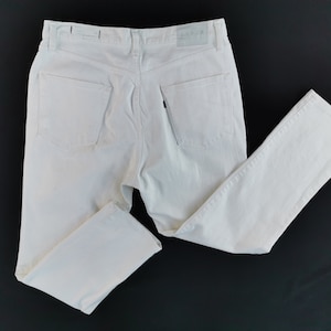 May include: Off-white denim jeans with a classic five-pocket design. The jeans have a straight leg cut and a button and zip closure. The back pockets are visible. The jeans are laid flat on a black surface.