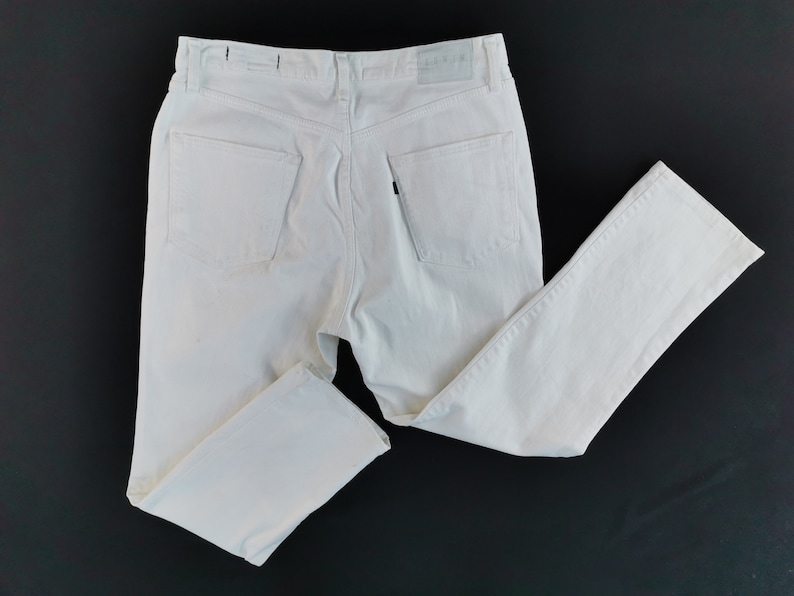 May include: Off-white denim jeans with a classic five-pocket design. The jeans have a straight leg cut and a button and zip closure. The back pockets are visible. The jeans are laid flat on a black surface.