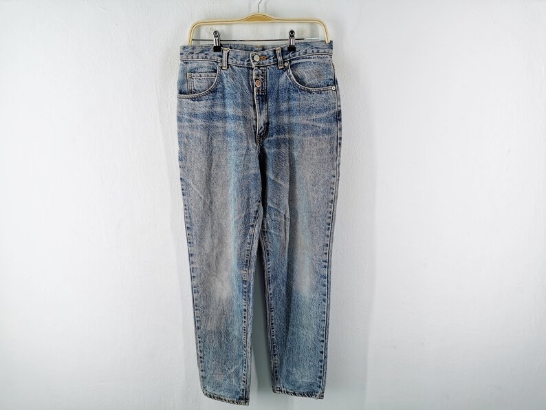 Op de afbeelding: Acid wash denim jeans met een knoopsluiting en een klassiek ontwerp met vijf zakken. De jeans hangen aan een gouden hanger tegen een witte achtergrond. De jeans hebben een vervaagde, versleten look.