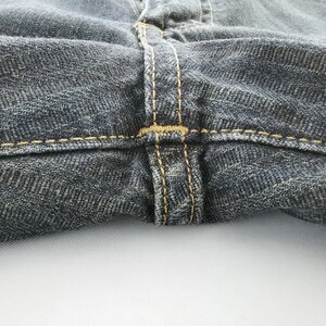 Peut inclure: Gros plan sur un jean en denim fonc&eacute;. L'image met en &eacute;vidence les d&eacute;tails des coutures autour de la ceinture et de la braguette. Le denim a un aspect textur&eacute;, avec des motifs de tissage diagonaux visibles. Les coutures sont de couleur beige contrastante.