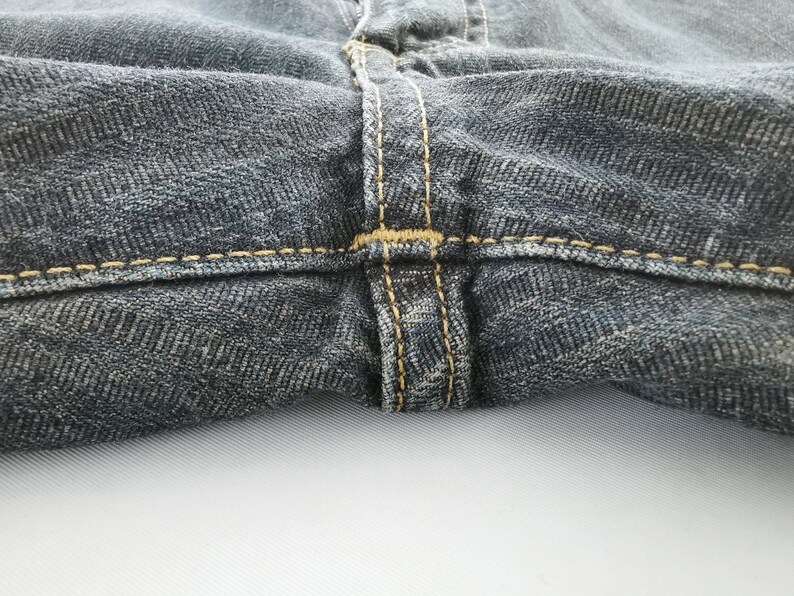 Peut inclure: Gros plan sur un jean en denim fonc&eacute;. L'image met en &eacute;vidence les d&eacute;tails des coutures autour de la ceinture et de la braguette. Le denim a un aspect textur&eacute;, avec des motifs de tissage diagonaux visibles. Les coutures sont de couleur beige contrastante.