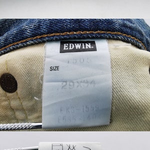 May include: Close-up of blue denim jeans with visible stitching and a white tag. The tag reads "EDWIN" and includes care instructions and fabric composition details. The jeans are made of 100% cotton.