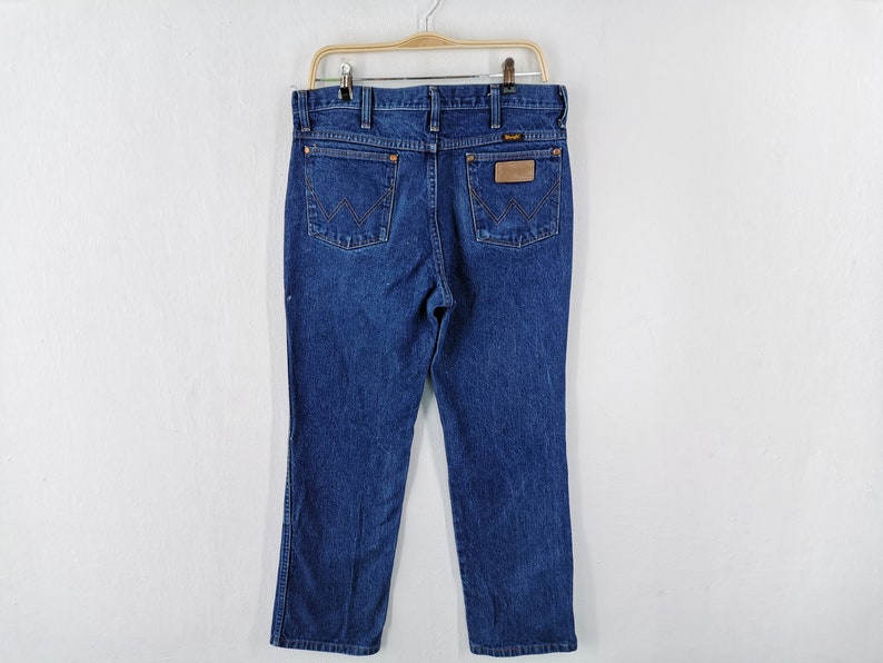 May include: Dark blue denim jeans hanging on a hanger. The jeans have a classic five-pocket design, a straight leg, and a brown leather patch with the brand name. The back pockets feature a distinctive stitched design.