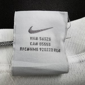 May include: A close-up shot of a white fabric label featuring the Nike swoosh logo. The label displays the text "RN# 56323", "CA# 05553", and "RFC#NME 920220 KL4". The label is attached to white ribbed fabric.