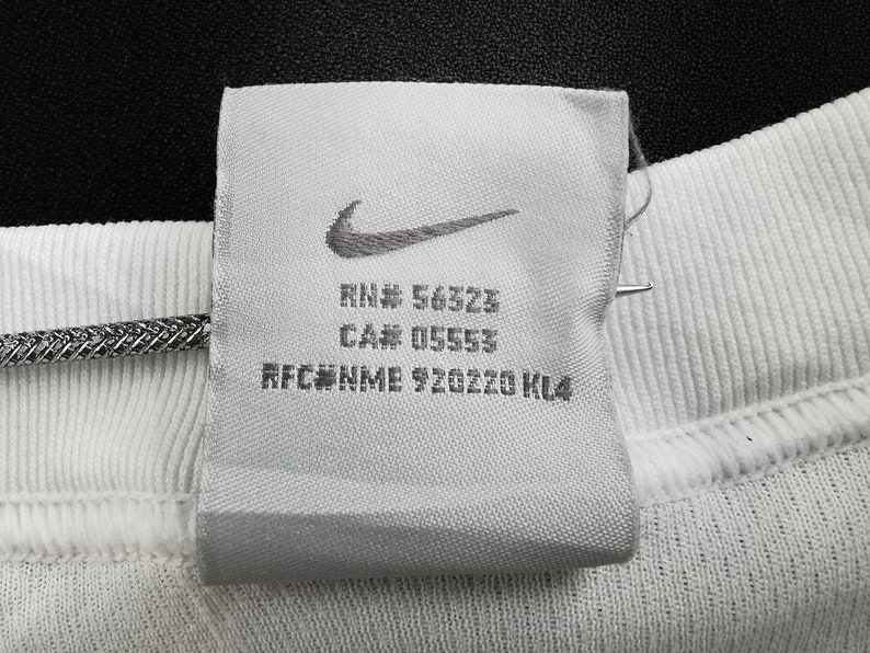 May include: A close-up shot of a white fabric label featuring the Nike swoosh logo. The label displays the text "RN# 56323", "CA# 05553", and "RFC#NME 920220 KL4". The label is attached to white ribbed fabric.