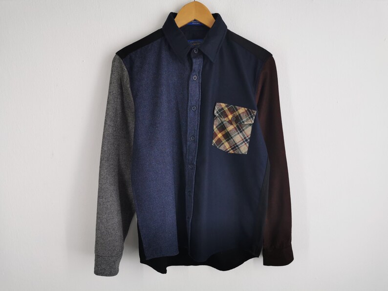 K&ouml;nnte beinhalten: Ein Button-Down-Hemd mit Patchwork-Design. Das Hemd hat einen marineblauen K&ouml;rper, eine denimartige Partie, einen grauen &Auml;rmel und einen weinroten &Auml;rmel. Eine karierte Tasche befindet sich auf der Brust. Das Hemd h&auml;ngt an einem Holzb&uuml;gel.