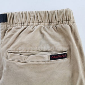 May include: Close-up of tan-colored pants with an elastic waistband and a black fabric loop. A small rectangular pocket is visible, with a red and black "GRAMICCI" label.