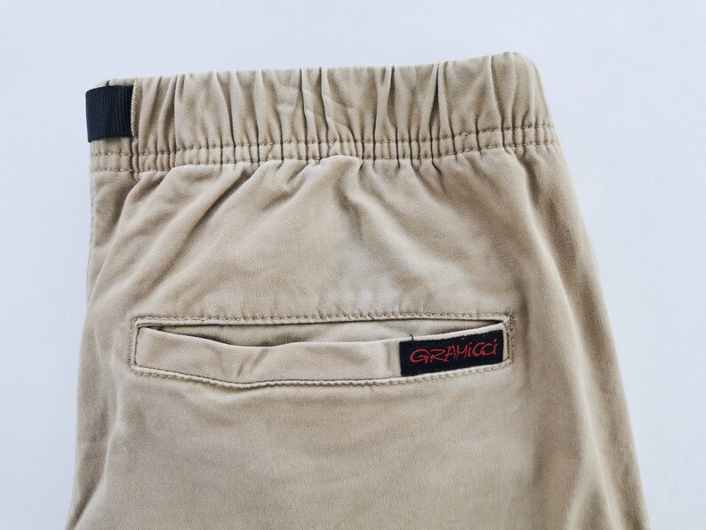 May include: Close-up of tan-colored pants with an elastic waistband and a black fabric loop. A small rectangular pocket is visible, with a red and black "GRAMICCI" label.