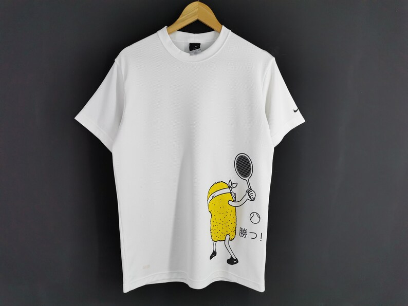 May include: White t-shirt with a cartoon tennis player graphic. The graphic features a yellow character with a headband holding a tennis racket, with a tennis ball and Japanese text. Nike logo on the sleeve.