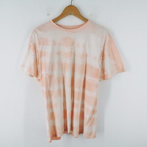 May include: A short-sleeved crew neck t-shirt featuring a horizontal tie-dye pattern in shades of soft peach and white. The garment is displayed on a wooden hanger, showcasing a relaxed fit with a round neckline and standard hemline construction.