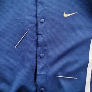 May include: A close-up of a blue jacket with a yellow Nike logo and snap closures. The jacket has white and yellow stripes on the sides. Two silver pins are placed on the jacket.